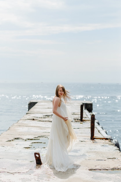 Een jonge mooie vrouw in een lange witte jurk loopt langs het strand en pier tegen de zee.