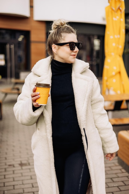Een jonge mooie vrouw in een witte bontjas en zonnebril met koffie loopt door de stad Stijlvol jong meisje