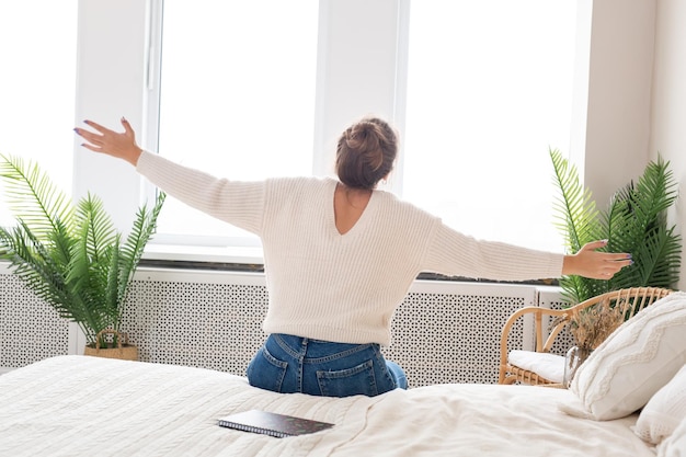 Een jonge mooie vrouw in een witte trui en spijkerbroek zit op het bed en kijkt uit het raam het meisje denkt en droomt over iets verheugt zich in de ochtend hoge kwaliteit foto