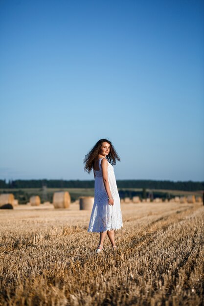 Een jonge mooie vrouw in een witte zomerjurk staat op een gemaaid tarweveld met enorme hooischoven, genietend van de natuur. Natuur in het dorp. Selectieve focus