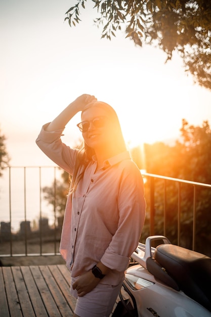 Een jonge mooie vrouw in zomerkleren staat op de achtergrond van een oranje zonsondergang in de buurt van de stadsbromfiets selectieve focus