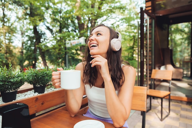 Een jonge mooie vrouw met koptelefoon zit aan een tafel in een zomercafé en drinkt koffie of thee generatie z