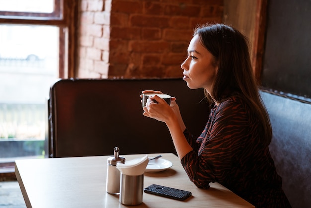 Foto een jonge mooie vrouw zit aan een tafel met een mok koffie bij het raam in een café, opzij kijkend met een peinzende blik