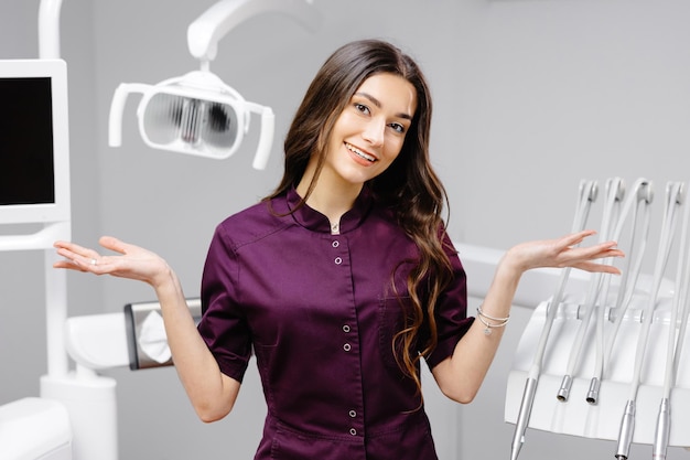 Een jonge mooie vrouwelijke tandarts staat in de buurt van de tandartsstoel op kantoor en gebaart met haar handen