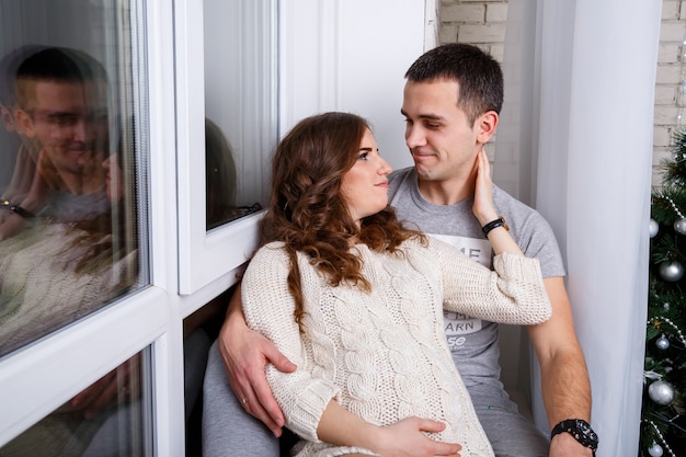 Een jonge, mooie zwangere vrouw en haar man in een comfortabele witte warme gebreide jurk en sokken zitten bij het raam, hand in hand op hun buik. Wit en grijs monochroom kleurenschema.