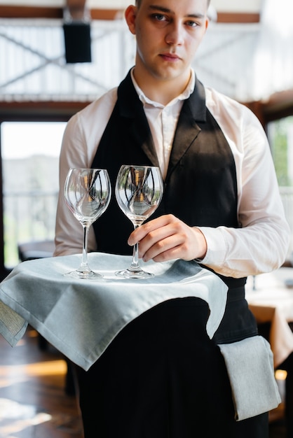 Een jonge ober in een stijlvol uniform staat met een bril op een dienblad bij de tafel in een prachtig gastronomisch restaurant close-up. Restaurantactiviteit, van het hoogste niveau.