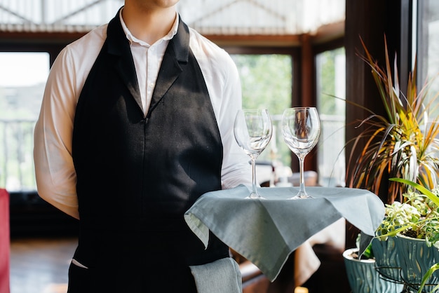 Een jonge ober in een stijlvol uniform staat met een bril op een dienblad bij de tafel in een prachtig gastronomisch restaurant close-up. Restaurantactiviteit, van het hoogste niveau.