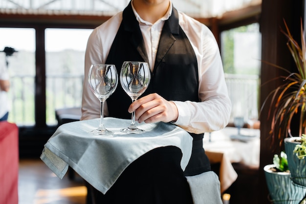 Een jonge ober in een stijlvol uniform staat met een bril op een dienblad bij de tafel in een prachtig gastronomisch restaurant close-up. Restaurantactiviteit, van het hoogste niveau.