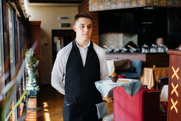 Een jonge ober in een stijlvol uniform staat met een voortreffelijk gerecht op een dienblad bij de tafel in een prachtig restaurant close-up. Restaurantactiviteit, van het hoogste niveau.