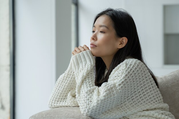 Een jonge ongelukkige aziatische vrouw die op de bank zit met een droevig gezicht en uit het raam kijkt