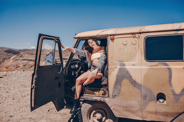 Een jonge prachtige vrouw poseren zittend in een vintage auto in de open natuur. Achter de heuvels en heldere wolkenloze lucht. Een reis op reis.