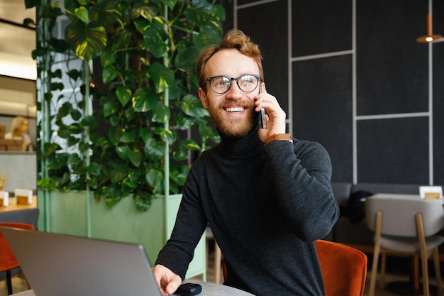 Foto een jonge roodharige man, een programmeur of ondernemer met een bril, zit in een stijlvol café achter een laptop en spreekt aan de telefoon freelancer werkt op afstand online communicatie kleine bedrijven