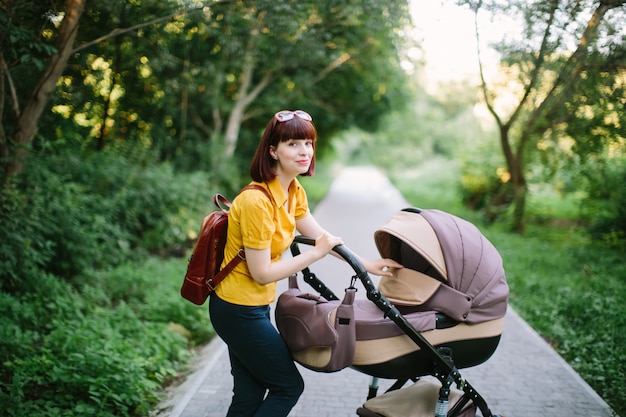 Een jonge roodharige vrouw in een geel shirt loopt met een kleine baby in een wandelwagen op een heldere zomerdag in het park