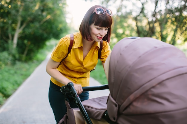 Een jonge roodharige vrouw in een geel shirt loopt met een kleine baby in een wandelwagen op een heldere zomerdag in het park