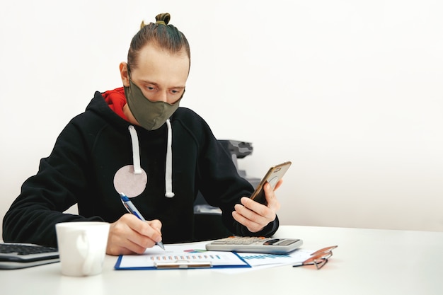 Een jonge specialist in een beschermend masker werkt met financiële documenten op een computer