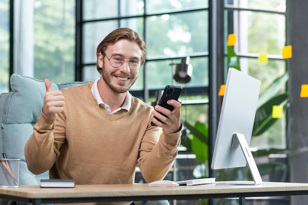 Een jonge student freelancer-programmeur die vanuit huis werkt, zit aan het bureau en gebruikt de telefoon om te schrijven