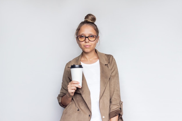 Een jonge succesvolle blonde vrouw in een bruine jas en bril houdt een kopje koffie of thee in wit papier