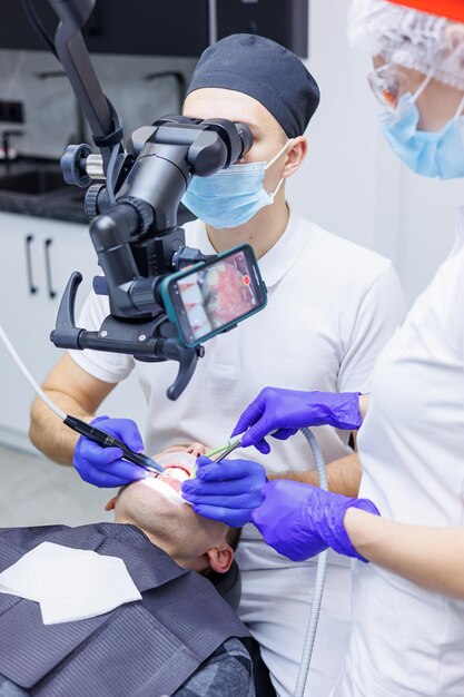 Een jonge tandarts kijkt naar de tanden van de patiënt met een tandheelkundige microscoop en houdt tandheelkundige instrumenten bij de mond een assistent helpt de dokter ze dragen witte uniformen met maskers