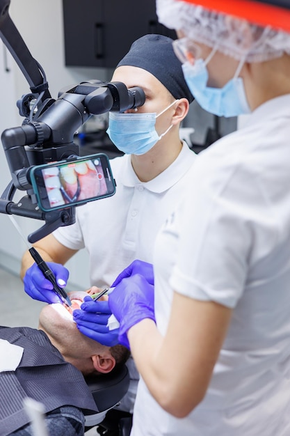 Een jonge tandarts kijkt naar de tanden van de patiënt met een tandheelkundige microscoop en houdt tandheelkundige instrumenten bij de mond Een assistent helpt de dokter Ze dragen witte uniformen met maskers