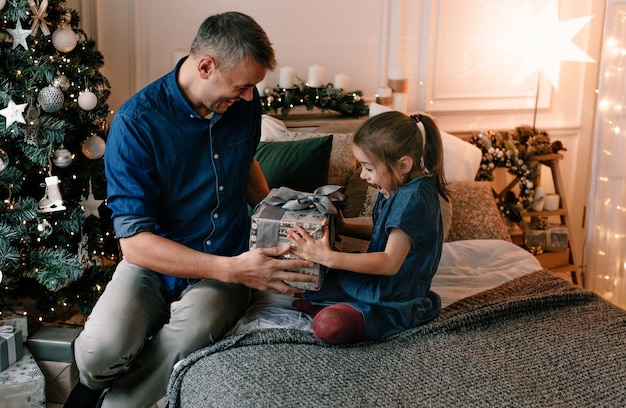 Een jonge vader geeft een kerstcadeau aan zijn lachende dochter bij een versierde kerstboom