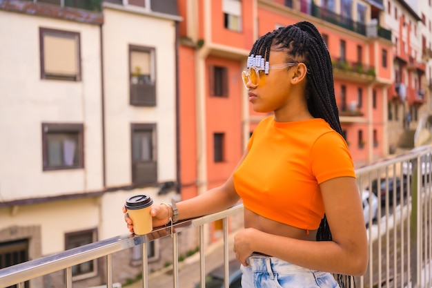 Een jonge valdanseres met vlechten. Zwart etnisch meisje met oranje t-shirt en cowboybroek met kopje Take-Away in the city