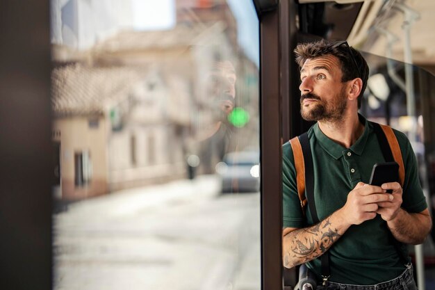 Foto een jonge verbaasde man reist in een stadsbus en kijkt door het raam terwijl hij zijn telefoon gebruikt