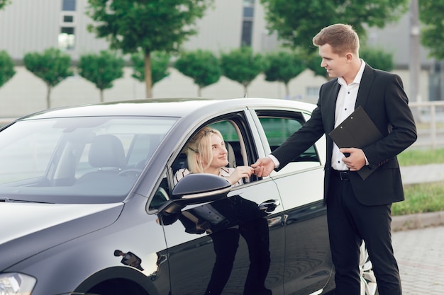 Een jonge verkoper laat klanten een nieuwe auto zien. Gelukkige vrouw koopt een nieuwe auto. De jonge vrouw zit achter het stuur, de verkoper geeft haar de sleutels.