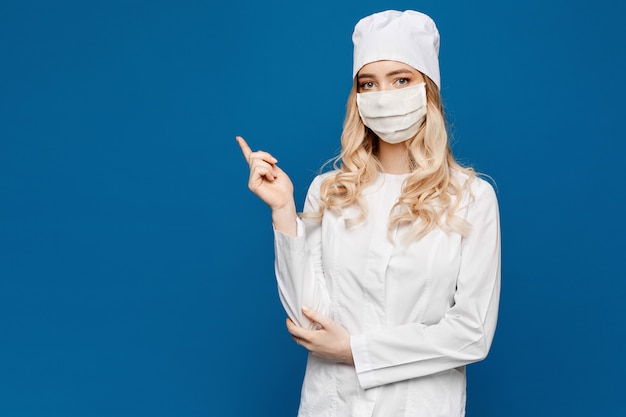 Een jonge verpleegster in een witte jas en medische masker vormt op een blauw en wijst een vinger naar de linkerkant, geïsoleerd met kopie ruimte. Jonge vrouw in medische uniform