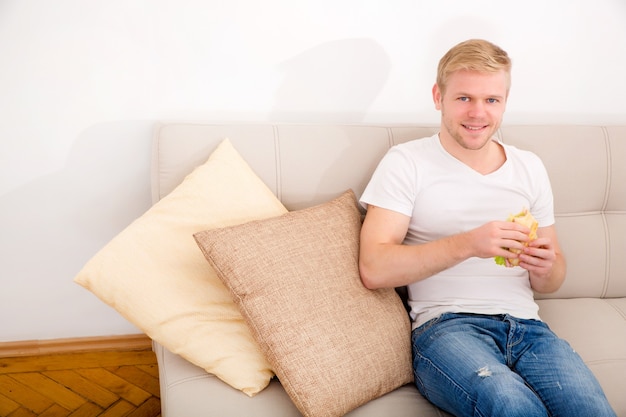 Een jonge volwassen man eet thuis een boterham op de bank.