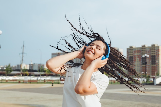 Een jonge vrolijke gelukkige vrouw met dreadlocks gekleed in een witte tshirt dansen luisteren naar muziek met koptelefoon rusten ontspannen in een stadspark wandelen langs een steegje Urban lifestyle concept