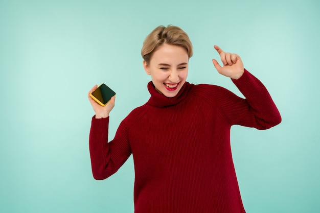 Een jonge vrolijke vrouw in een rode trui met een smartphone op een blauwe achtergrond