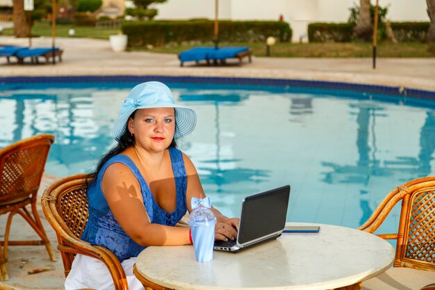 Een jonge vrouw aan een tafel in de ochtend naast het zwembad werkt op afstand op een laptop