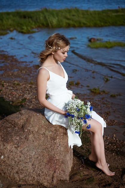 Een jonge vrouw alleen in een witte jurk zit op een steen aan de kust met een boeket in haar hand in de zomer bij zonnig weer