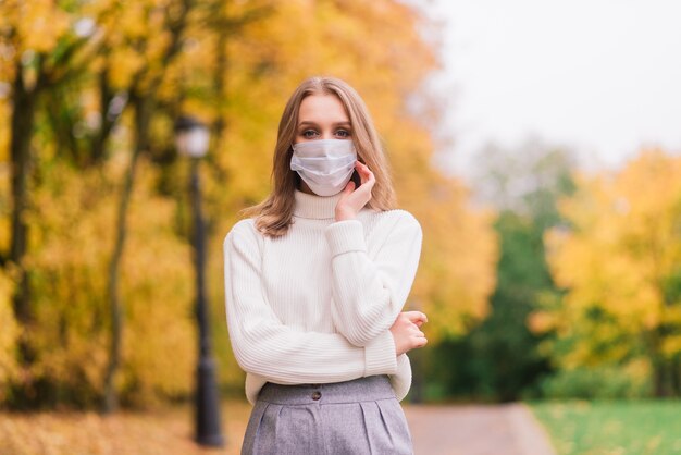 Een jonge vrouw die beschermt tegen het coronavirus tijdens het wandelen in het park. herfst achtergrond.
