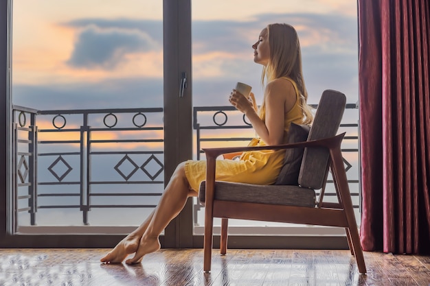 Een jonge vrouw die een koffiekopje vasthoudt terwijl ze op een stoel op het balkon zit en naar bergen en groen kijkt