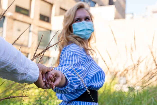 Foto een jonge vrouw die het beschermende masker draagt, neemt haar man bij de hand, nodigt hem uit om door de planten naar een helderdere ruimte te lopen, een conceptueel beeld van optimisme, focus op handen