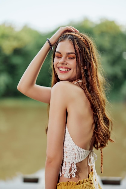 Een jonge vrouw die lacht in een afbeelding van een hippie en ecodress dansend in de natuur bij het meer met een hoed en een gele broek op in de zomerzonsondergang