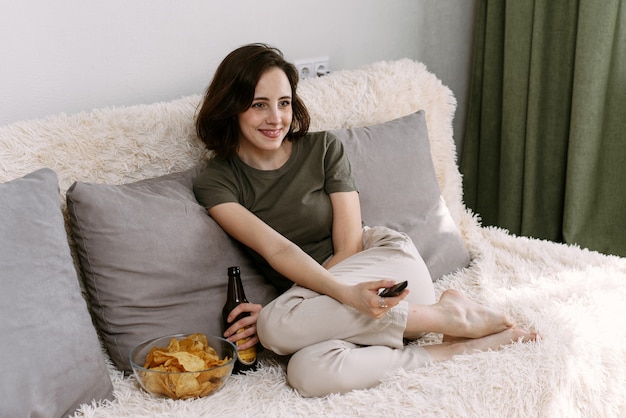 Een jonge vrouw drinkt bier en eet thuis chips. Thuis ontspannen tv en films kijken.