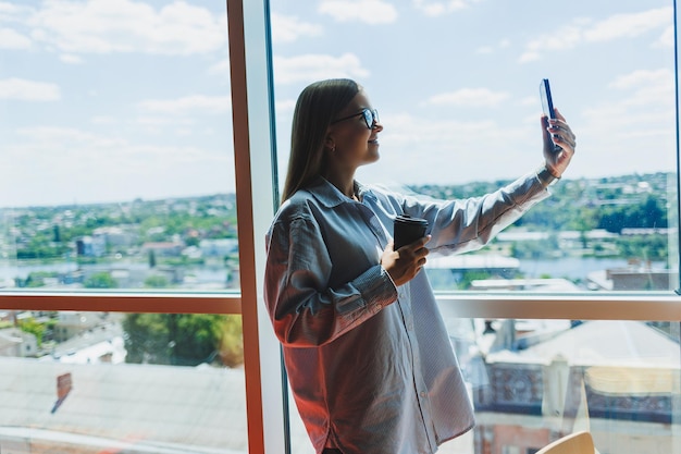 Een jonge vrouw freelancer met een bril staat met koffie en praat aan de telefoon bij een groot raam en kijkt naar de stad Een meisje in een shirt en korte broek staat in een café bij het raam