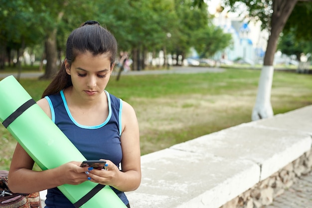 Een jonge vrouw houdt een yogamat vast en klimt aan de telefoon na een training op straat.