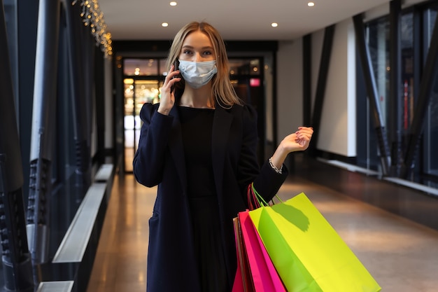 Een jonge vrouw in een beschermend masker na het winkelen praat aan de telefoon in een winkelcentrum