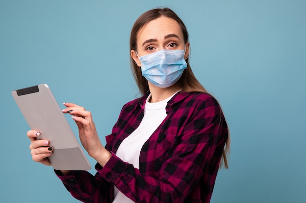 Een jonge vrouw in een beschermend masker typt op een tablet op een blauwe achtergrond