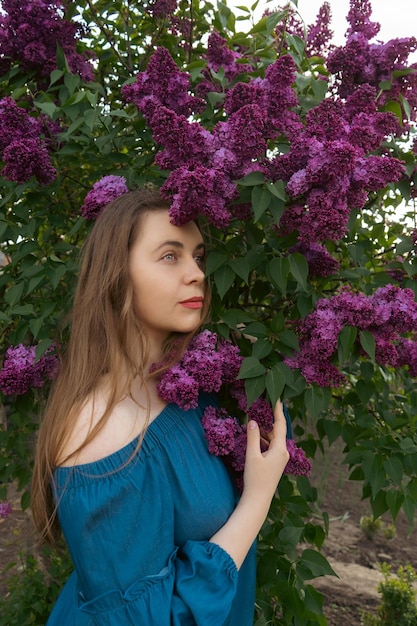 Een jonge vrouw in een blauwe jurk in een bloeiende lila tuin Lenteverhaal Bruinharige vrouw met lang haar Een vrouw speelt met haar haar Romantisch lentegezicht