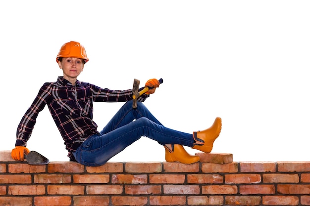 Een jonge vrouw in een bouwhelm met een bouwhamer en een troffel in haar handen zit op een bakstenen muur die op een witte achtergrond wordt geïsoleerd