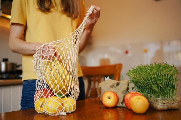 Foto een jonge vrouw in een gele t-shirt haalt appels en eten uit een netzak op tafel close-up concept van gezonde voeding, ecologische verpakkingen en biologische producten