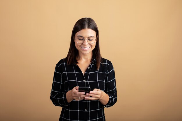 Een jonge vrouw in een geruite jurk staat op een oranje achtergrond en speelt met haar telefoon