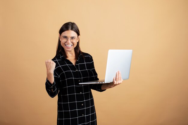 Een jonge vrouw in een geruite jurk staat op een oranje achtergrond met een laptop