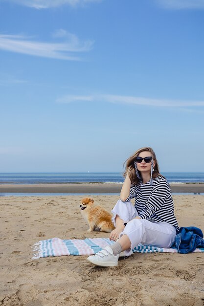 Een jonge vrouw in een gestreept shirt met een kleine Pommerse hond zittend op een deken op een zandstrand