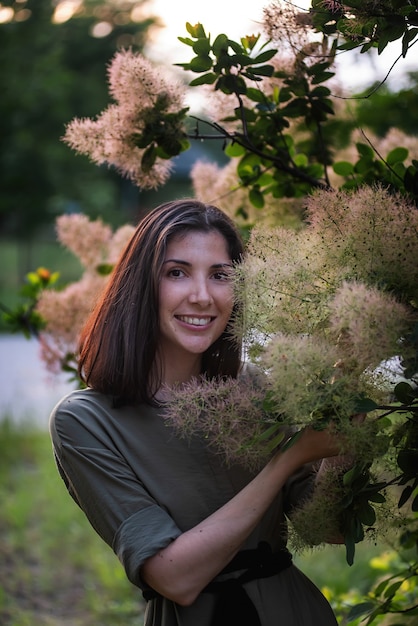 Een jonge vrouw in een groene kaki jurk staat bij een rozenstruik smoky skumpia bij zonsondergang in het park