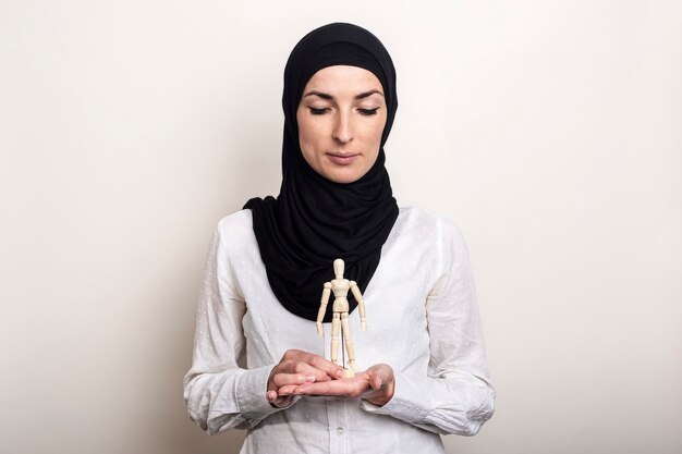 Een jonge vrouw in een hijab houdt kijkt naar een marionet van een houten man op een lichte achtergrond.